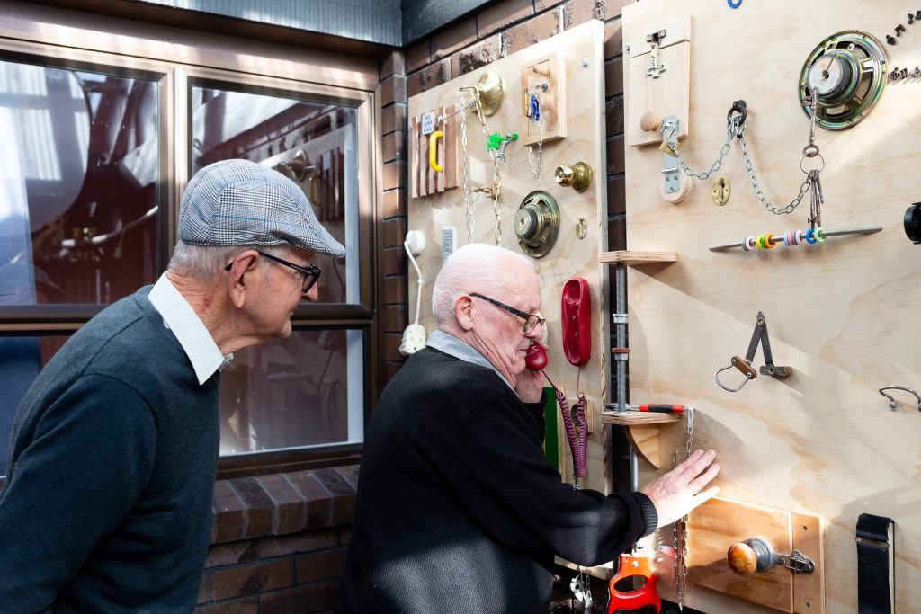 Two elderly men examine a wall of interactive gadgets, tools and equipment.