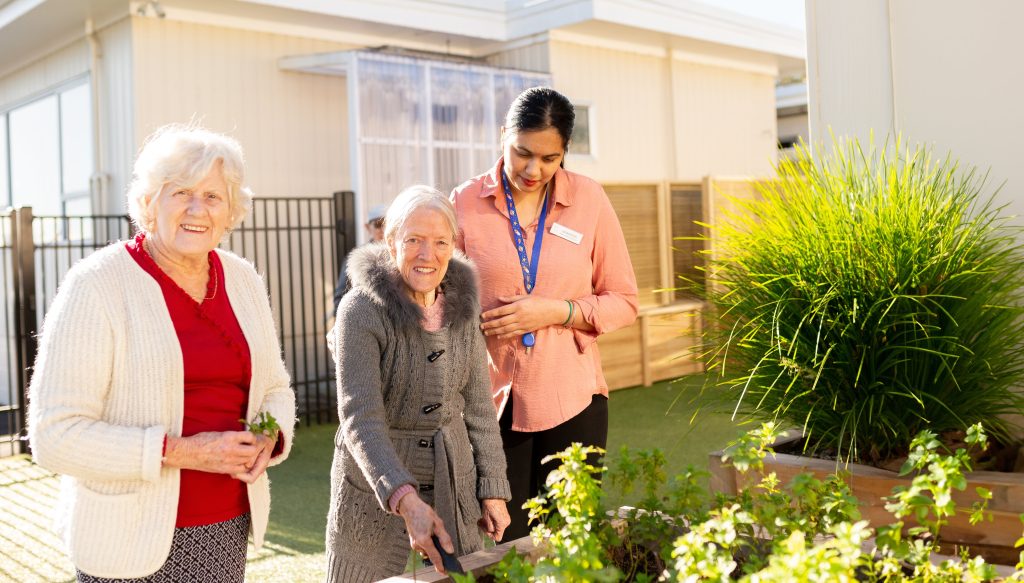 A carer helps two elderly ladies garden in a planter box. Gardening is one fo the great dementia friendly summer activities